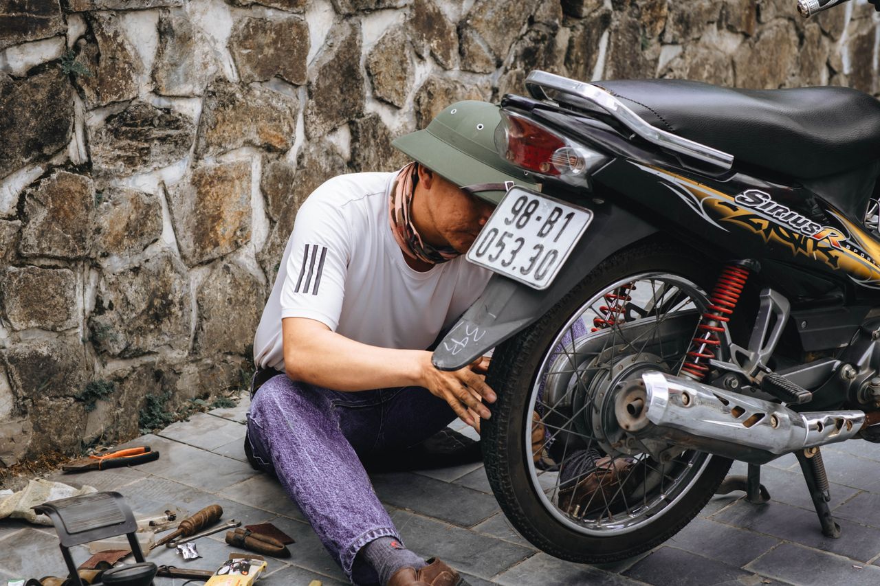 A mechanic working outdoors repairing a motorbike rear wheel with tools scattered around.