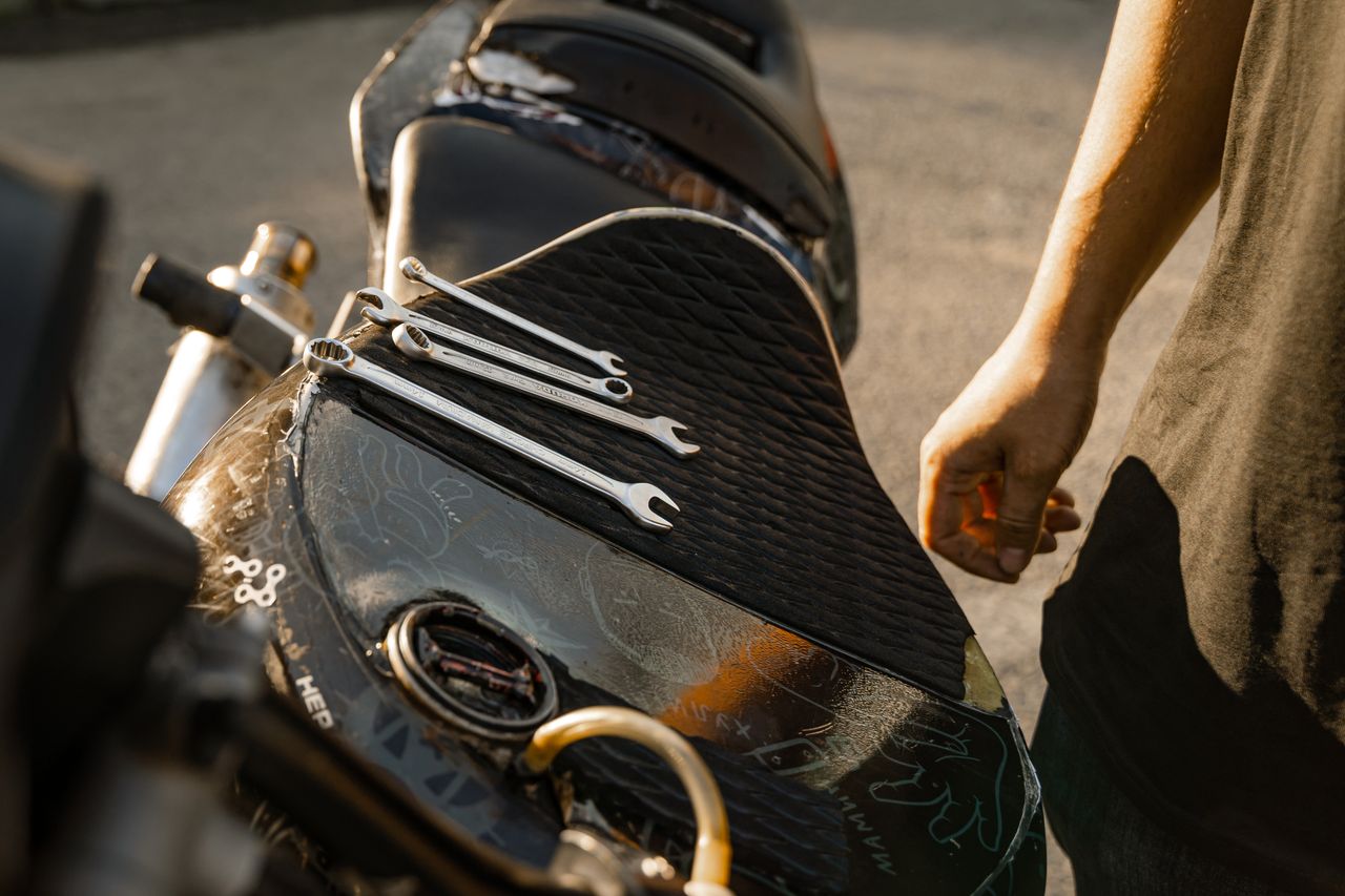 Close-up of motorcycle seat with wrenches, hand nearby. Outdoor setting, bright sunlight.